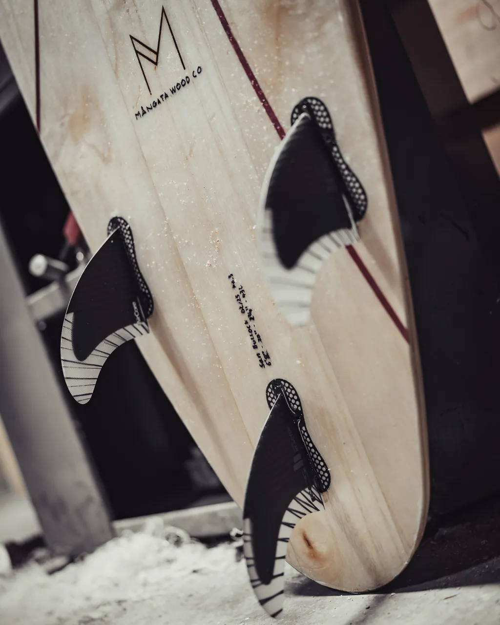 Close view of a board's tail and fins, the grain of the timber and two oxblood lines curving along the rails.