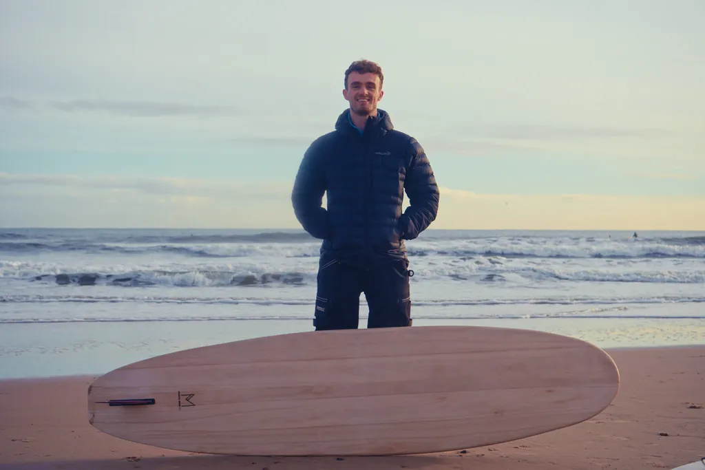 Sam stands on the beach in a winter jacket, smiling, with one of his wooden boards resting in front of him.