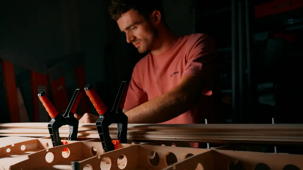 Sam works at the bench behind a row of clamps, a surfboard frame taking shape in front of him.