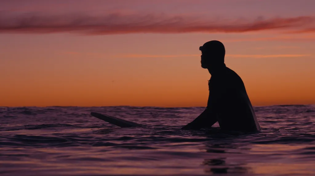 A surfer sits on a wooden board in the sea, silhouetted against an orange sunset sky.